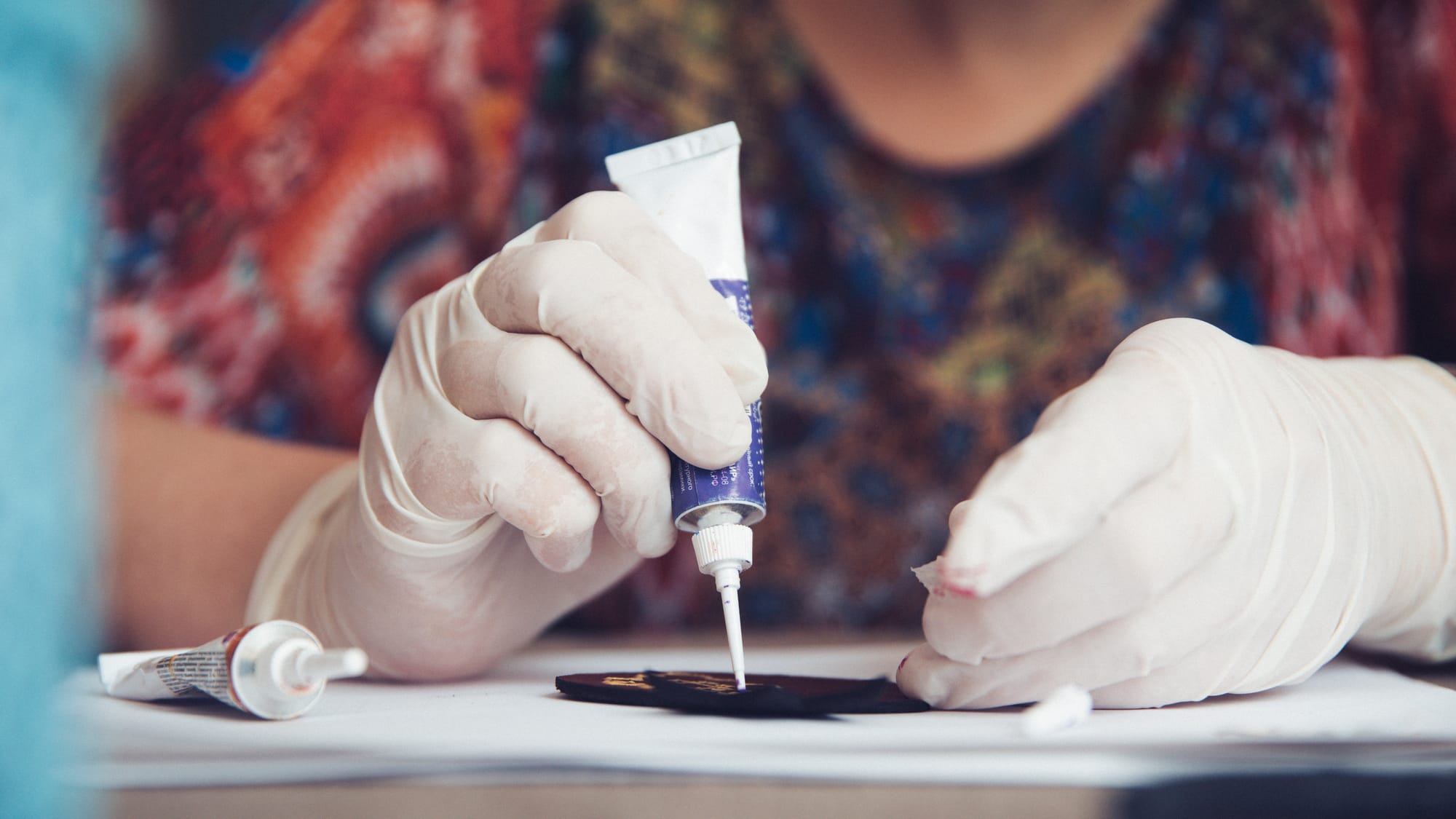 A close shot of an older woman in a colorful shirt wearing white protective gloves while using superglue