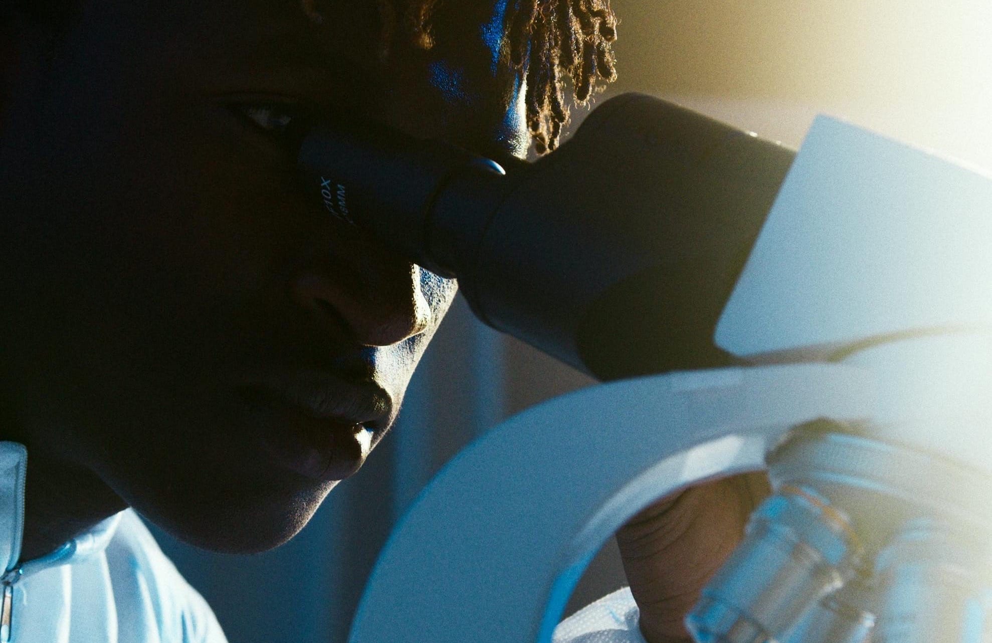 A stock photo of a Black male scientist with thin, ginger dreadlocks looking through a microscope